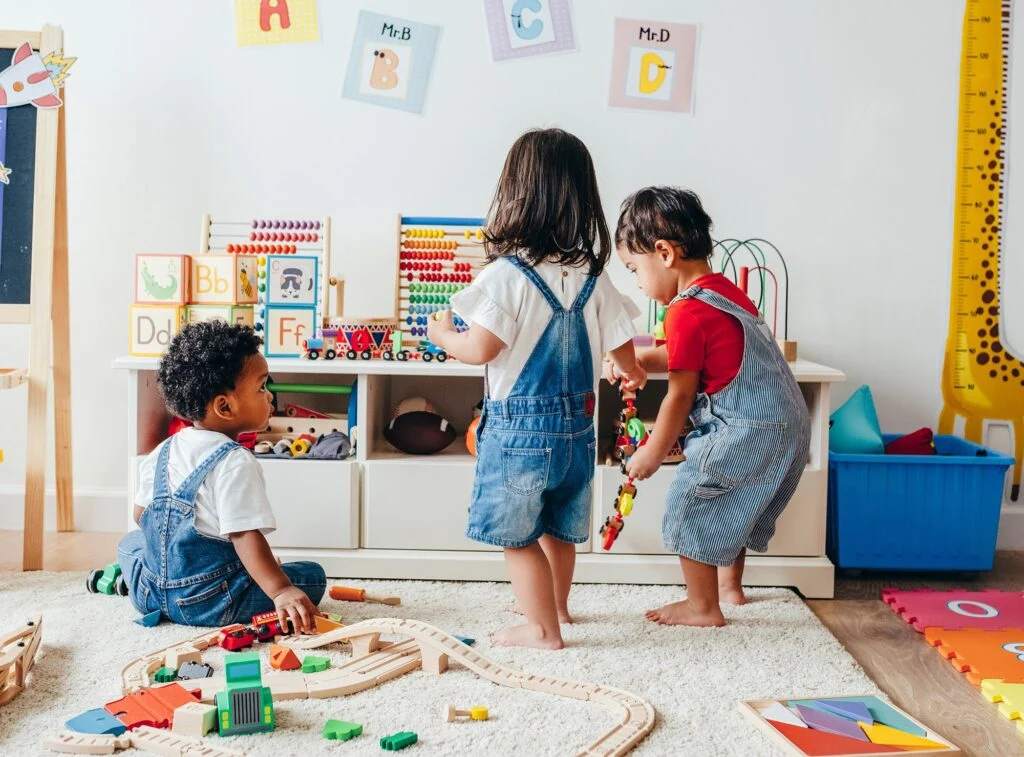 Two toddlers in denim overalls playing with toys and a wooden train in a bright playroom